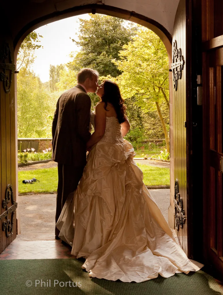 A couple stands embracing at the threshold of an arched doorway with a garden visible beyond.