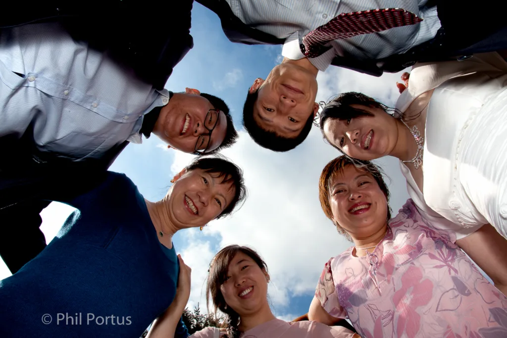 Six people smile upwards towards the camera against a bright blue sky with scattered clouds.