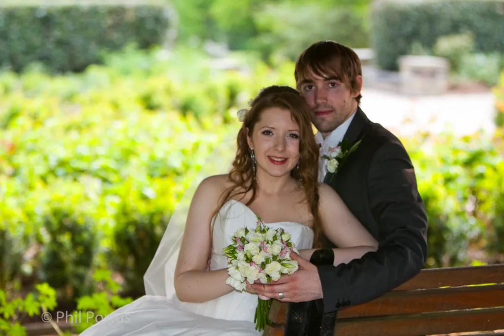 A couple dressed for a wedding sits on a wooden bench in front of lush greenery.