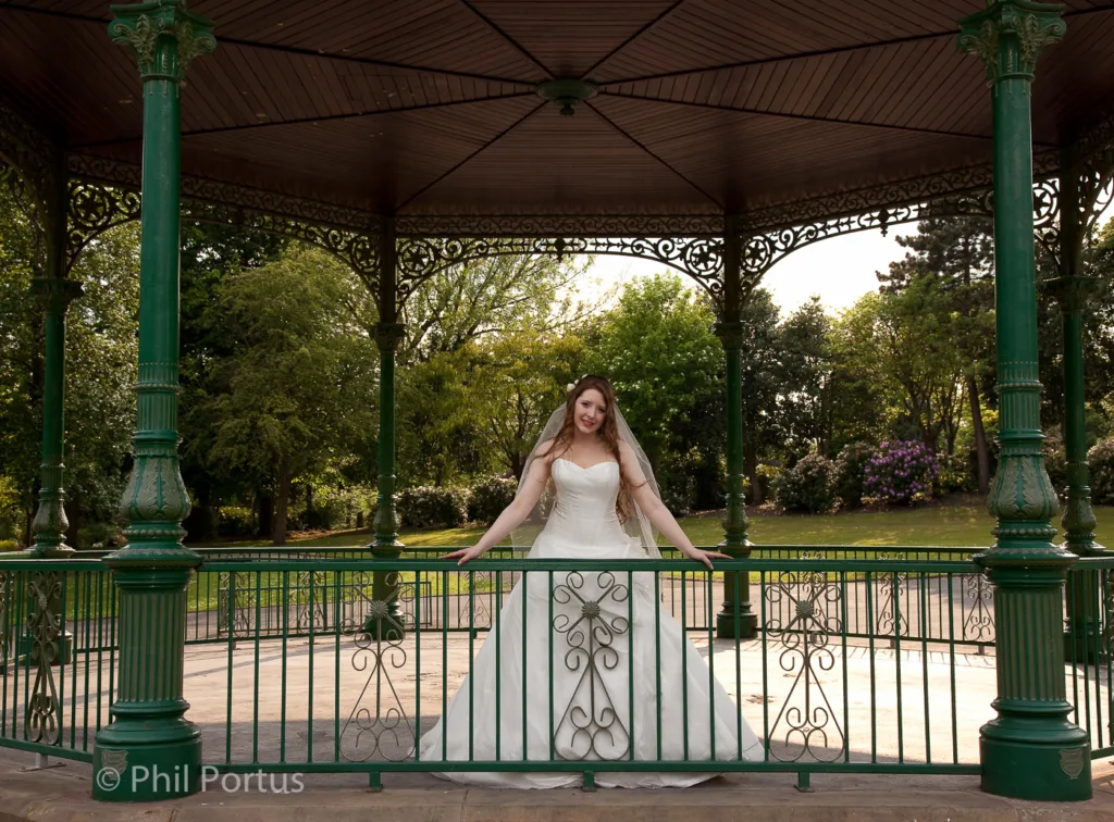 A person in a wedding dress leans on the railing of an ornate gazebo with greenery visible behind.