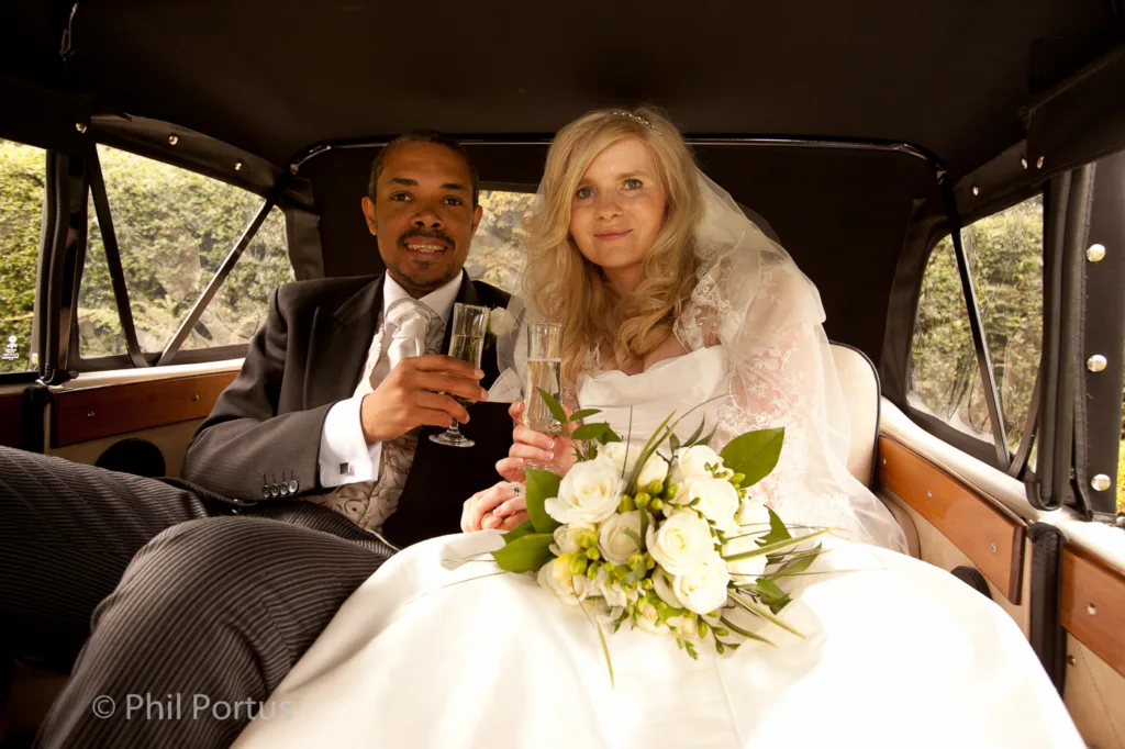 A couple in formal wedding attire sits inside a vintage car, raising glasses and smiling at the camera.