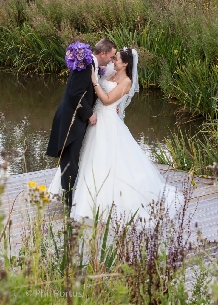 A couple in formal wedding attire embrace on a wooden boardwalk beside still water and tall grasses.