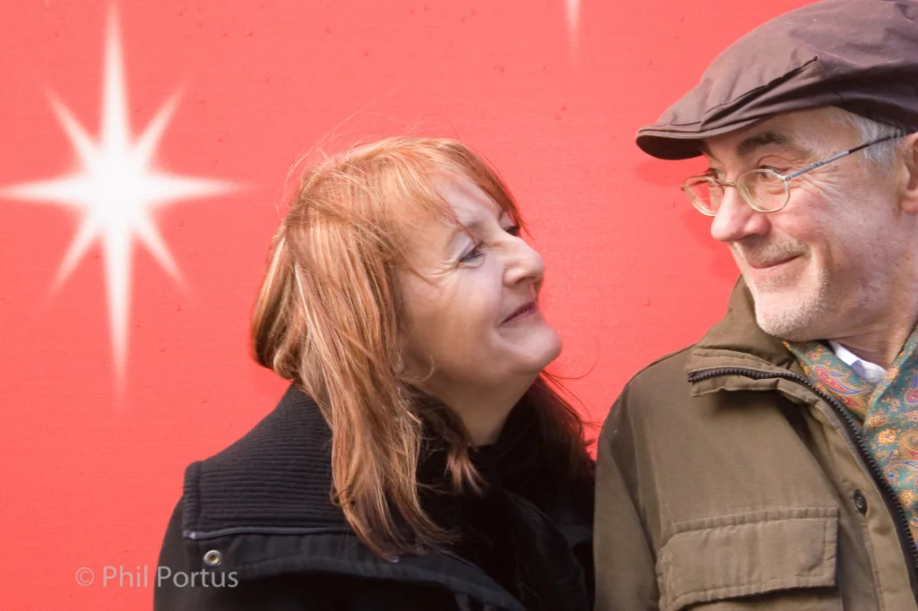 Two adults stand close together, looking at each other with smiles against a bright red backdrop.