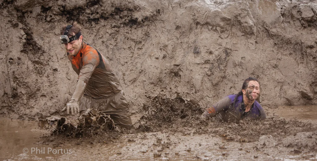 Two people struggle through thick mud, arms outstretched, with expressions of exertion.