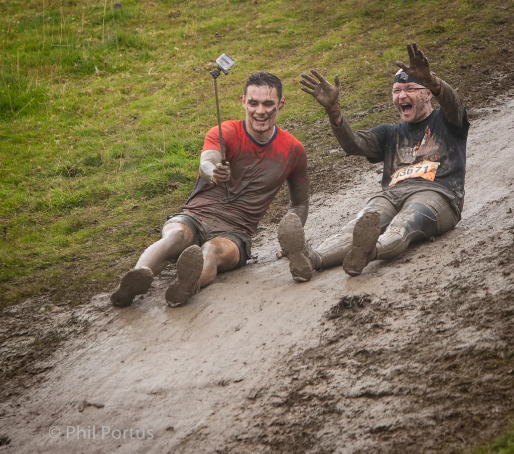 Two people covered in mud slide down a muddy slope, smiling and waving.