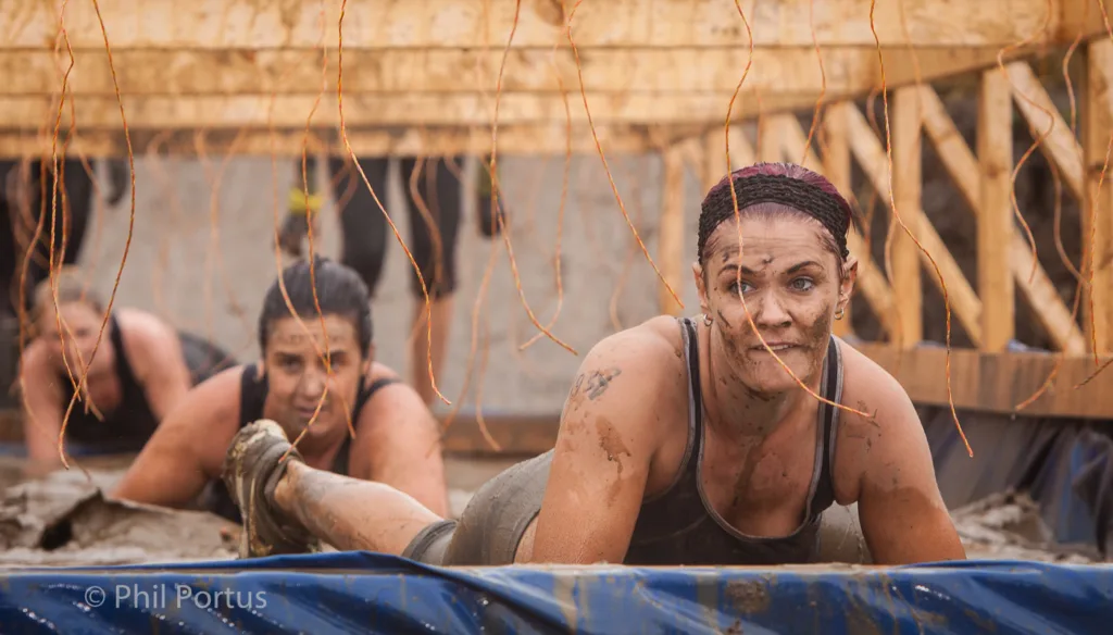Two people crawl under a low barrier covered in string during an obstacle course event.