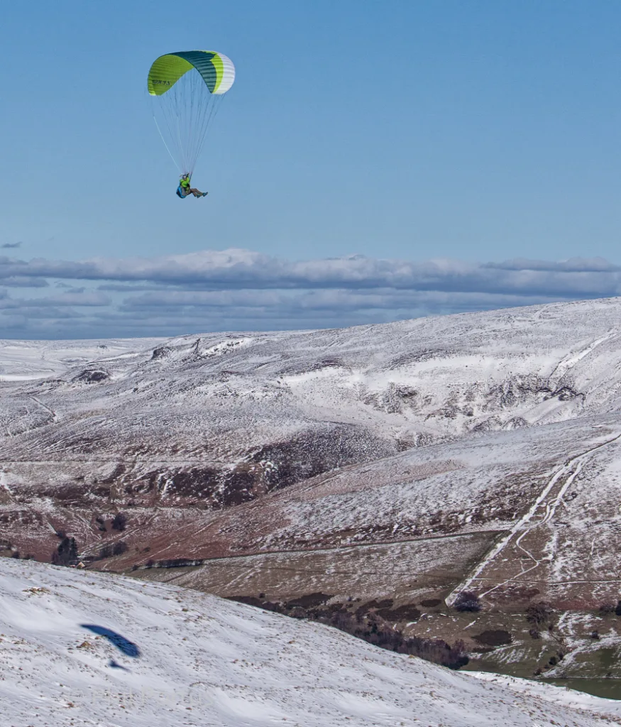 A paraglider flies over a snow-covered landscape under a clear blue sky.