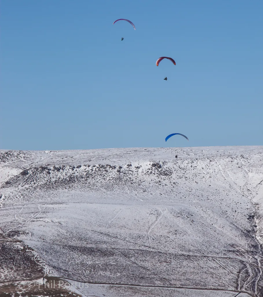 Three paragliders fly against a clear blue sky above a snow-covered hillside.
