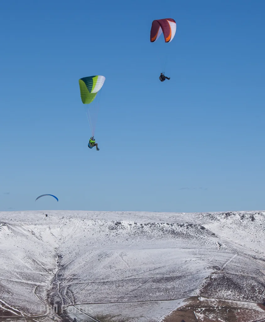 Three paragliders fly against a clear blue sky above a snow-covered landscape.