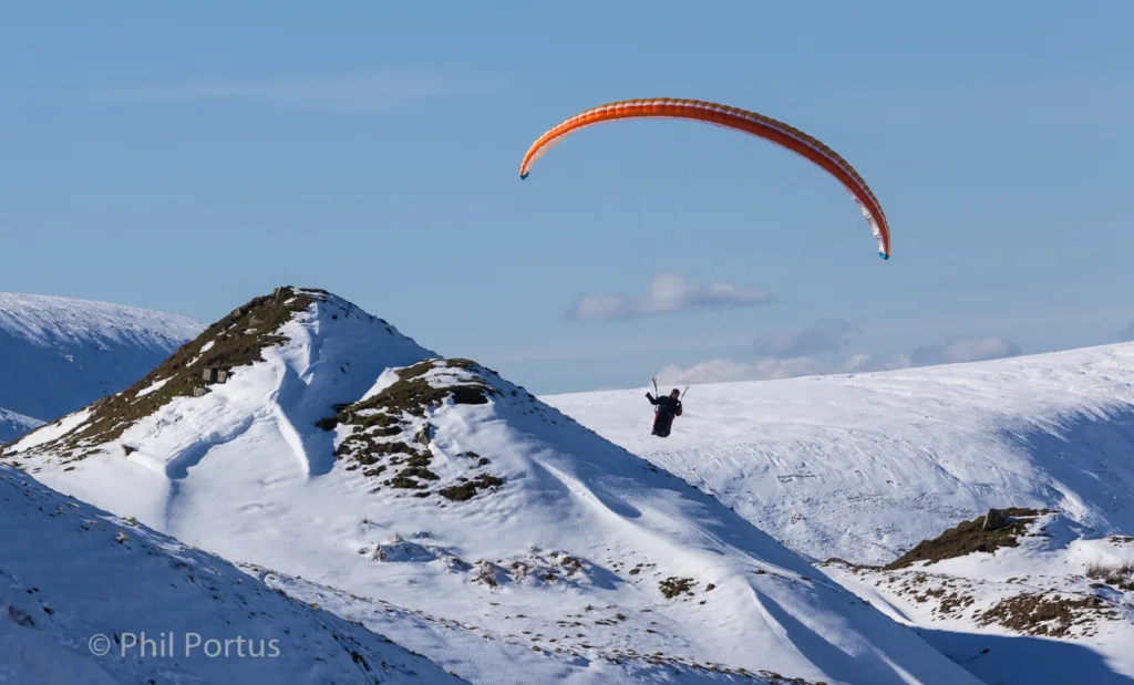 A paraglider flies over a snow-covered mountain range under a clear blue sky.