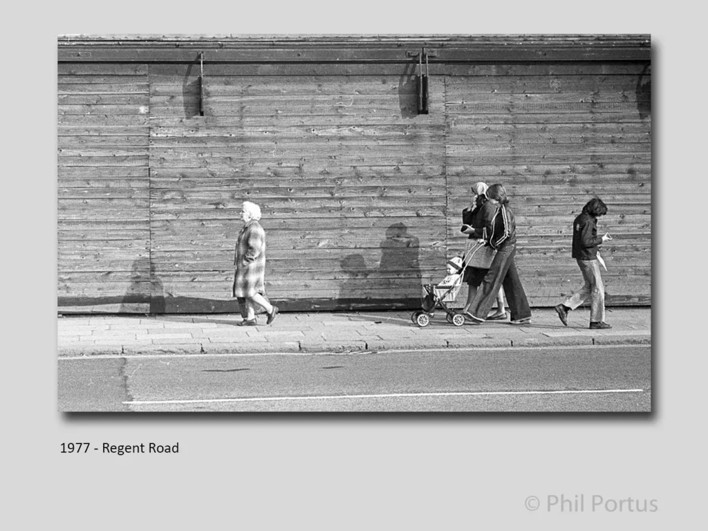 Three figures walk along a sidewalk next to a long wooden wall in black and white.