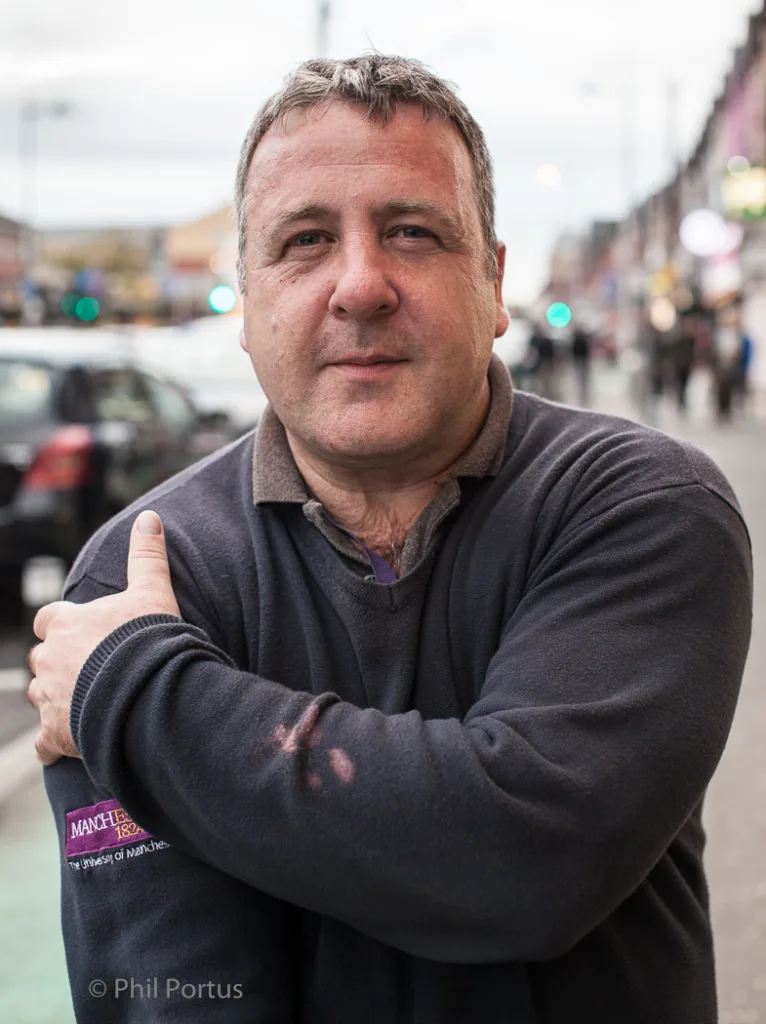 A middle-aged man with graying hair gives a thumbs up while standing on a street.