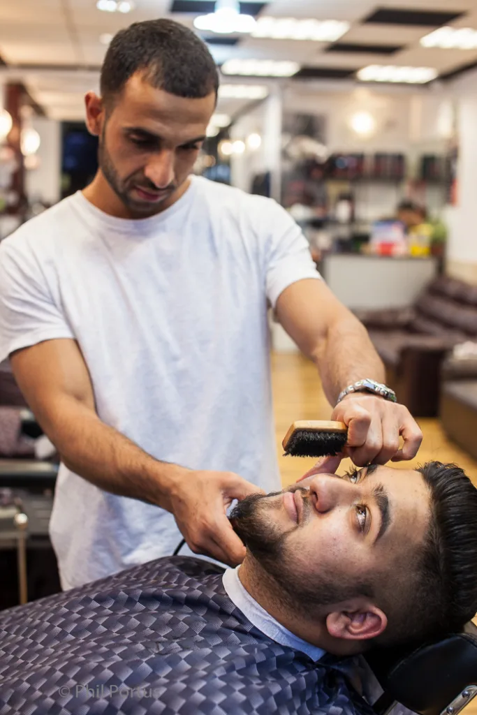 A barber uses a brush on a man's beard while he reclines in a salon chair.