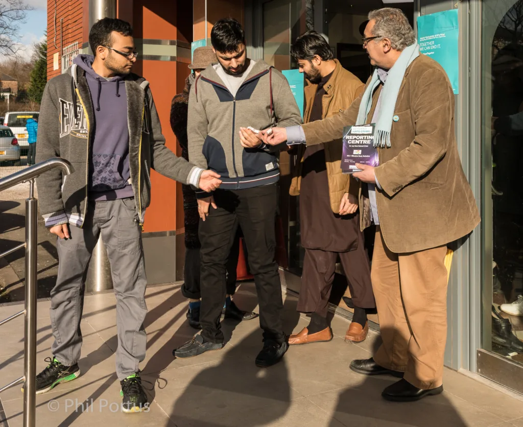 Four men stand outside a building on a sunny day, looking at a mobile phone.