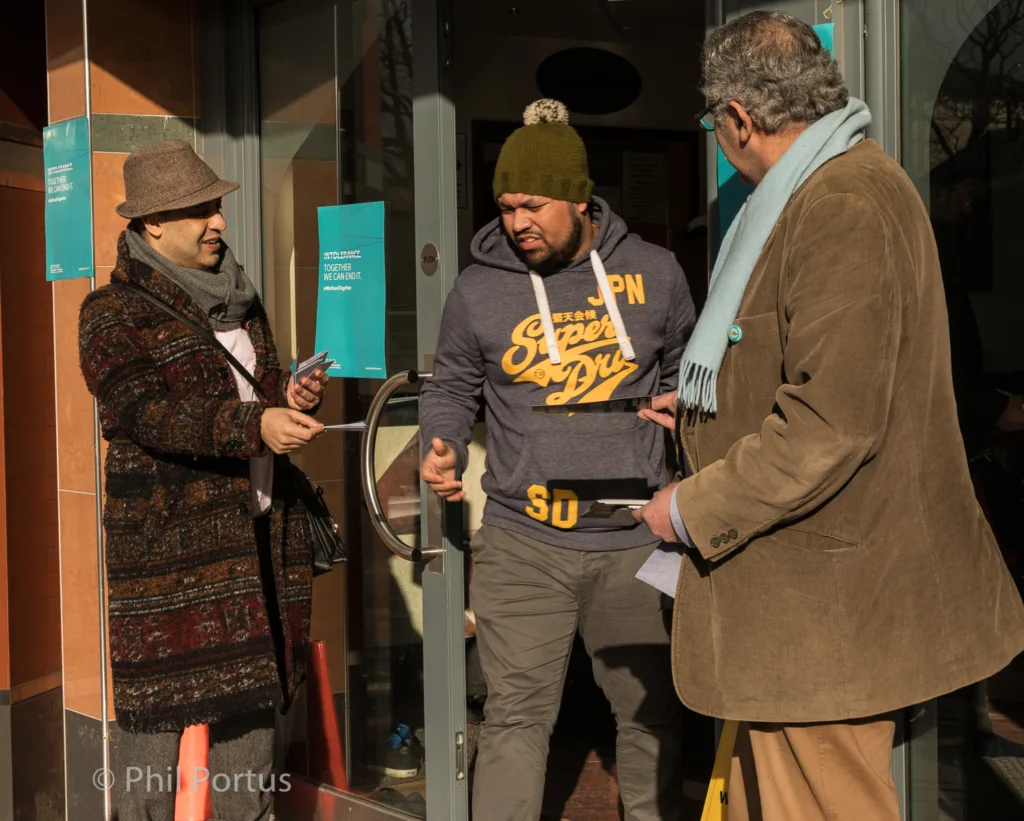 Three people stand near a glass door in bright sunlight, engaged in conversation.