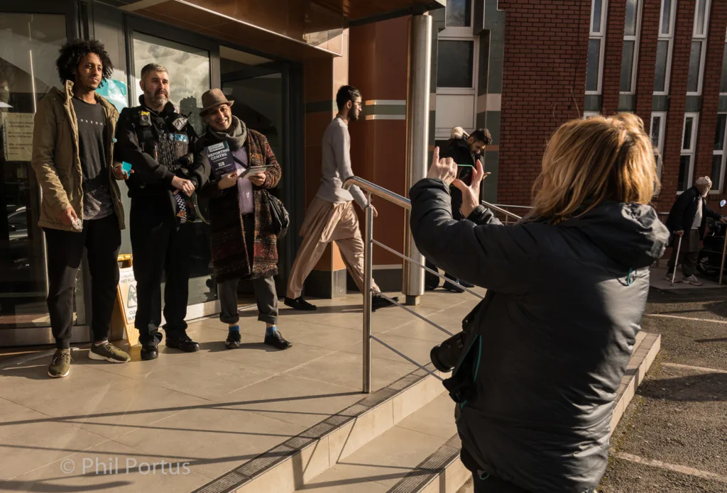 A person with short blonde hair takes a picture of several people standing near a building entrance.