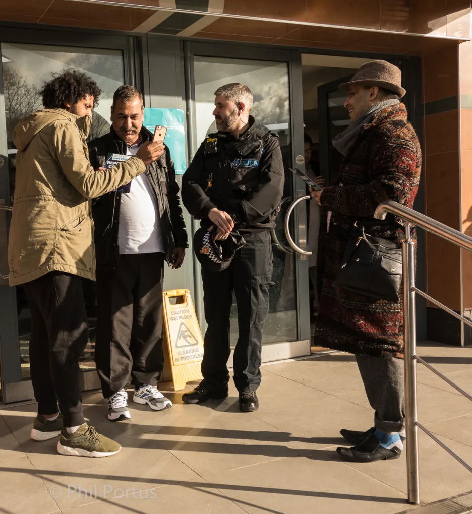 Four people stand near a glass entrance, two using mobile phones while a police officer observes.