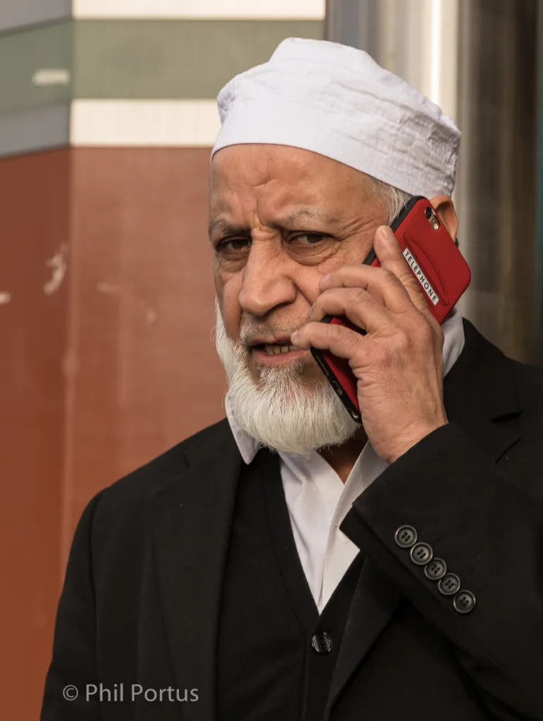 An older man with a white cap and beard speaks on a smartphone in a red case.