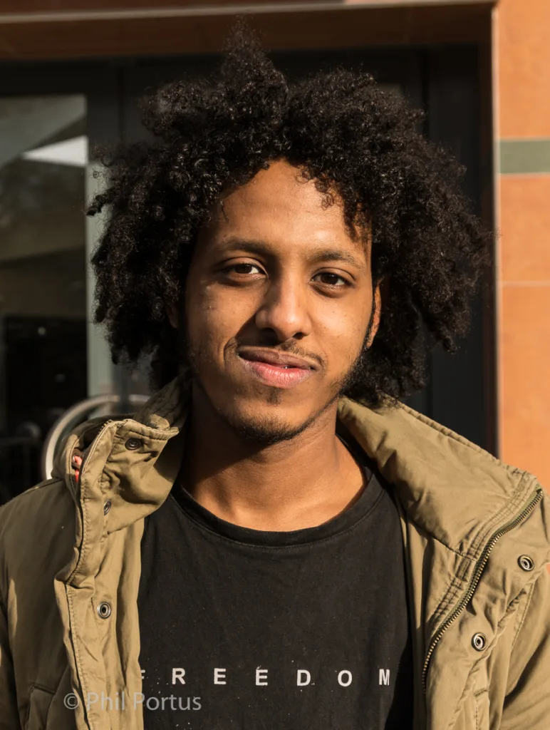A young man with curly dark hair looks directly at the camera wearing a black shirt and olive green jacket.