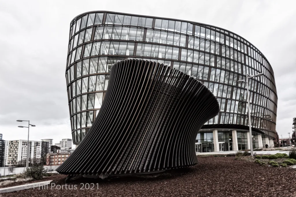 A modern curved building stands behind a large abstract sculpture made of dark metal strips.