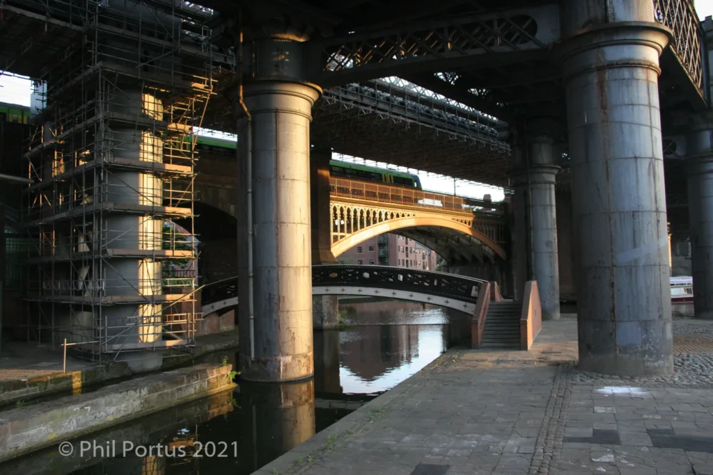 A canal runs between large stone pillars supporting a metal bridge structure with scaffolding nearby.
