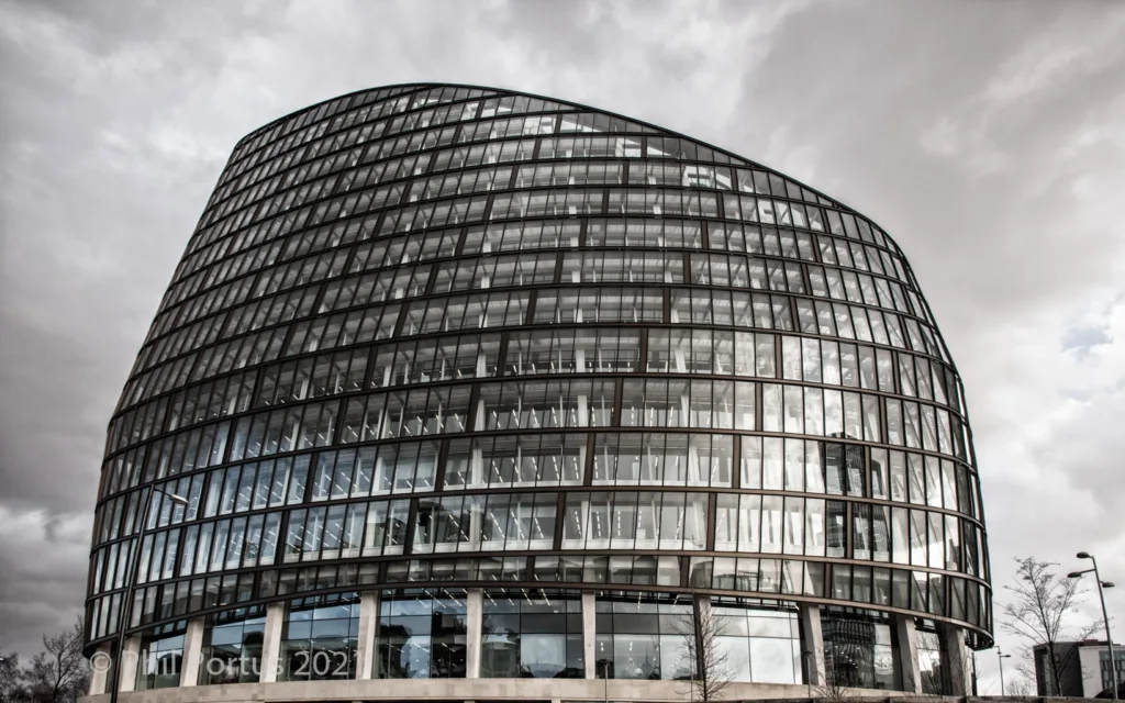 A tall, curved building with many windows rises against a cloudy sky.