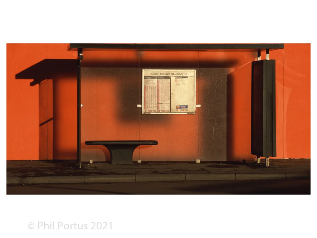 A bus shelter with a bench sits against an orange wall under strong directional light.