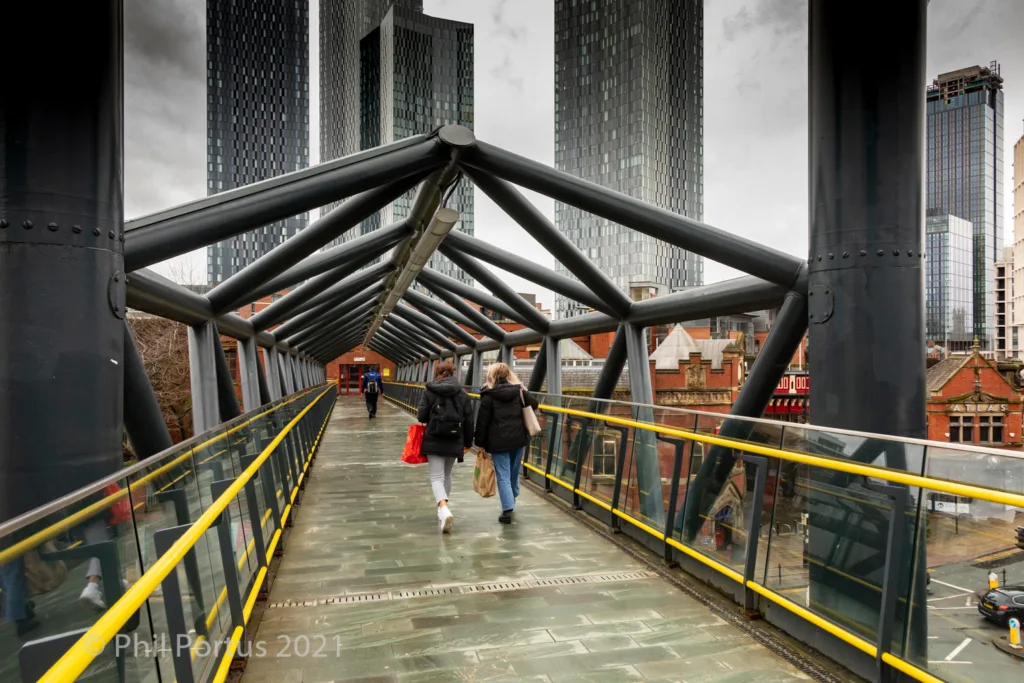 Two people walk along a covered pedestrian bridge with modern buildings rising in the background.