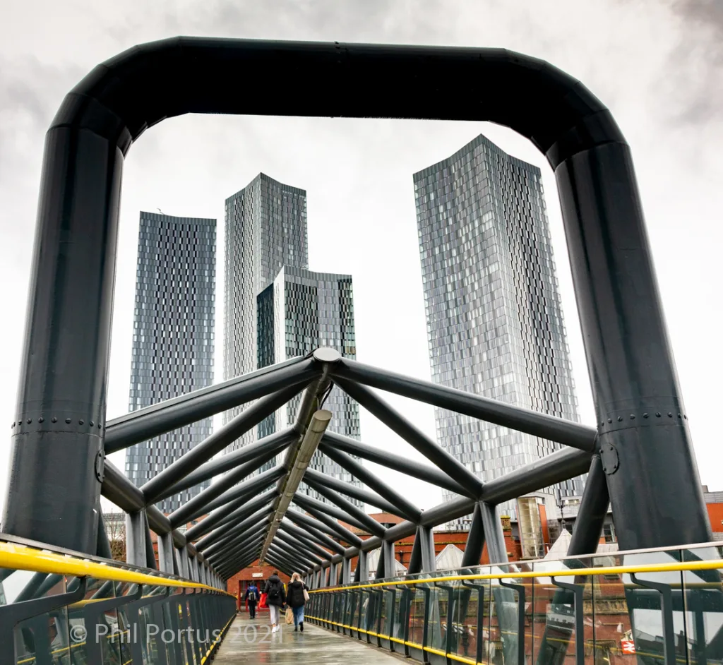 A black metal pedestrian bridge frames a view of three tall buildings with reflective glass facades.
