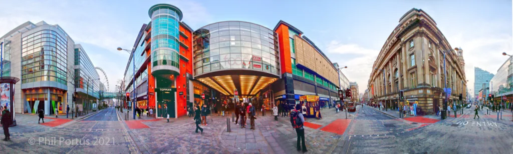 A wide panoramic view shows modern buildings with glass facades along a pedestrian street filled with people.