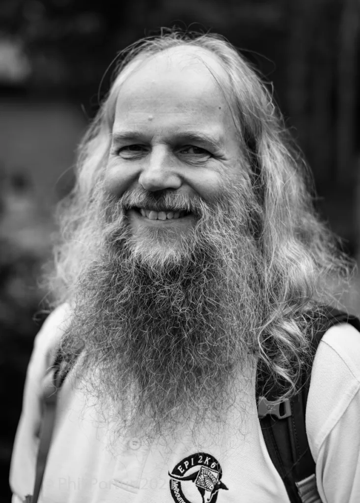 A man with a long beard and hair smiles directly at the camera in black and white.