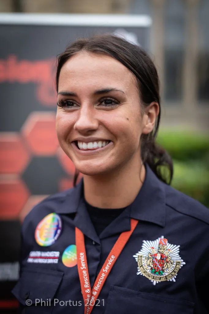 A smiling woman wearing a dark uniform shirt with ribbons and a badge is looking directly at the camera.