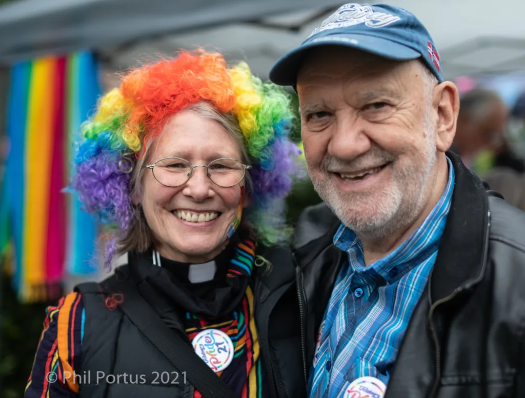Two people smile for a portrait; one wears a colorful wig and clerical collar.