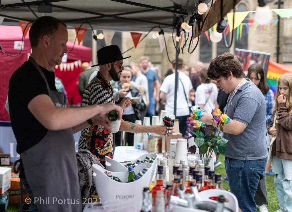 Three people interact at a bustling outdoor event under a dark canopy with tables displaying bottles and decorations.