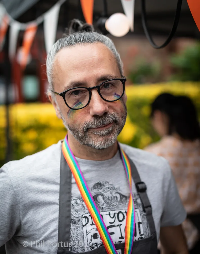 A person with graying hair pulled back in a bun looks directly at the camera while wearing glasses and an apron.