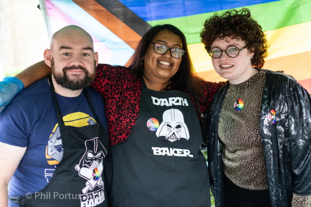 Three people smile for a portrait outdoors with a rainbow backdrop and colorful clothing.