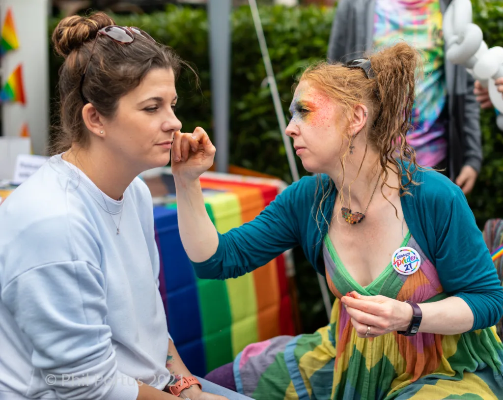 Two people sit close together outdoors; one applies face paint to the other's cheek.