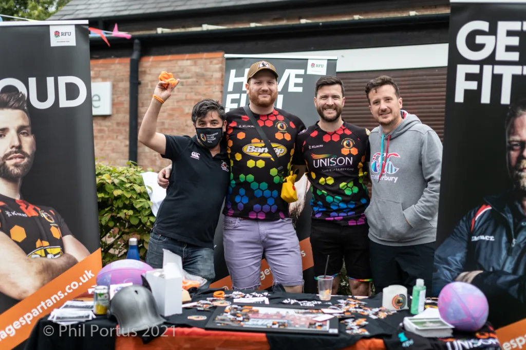 Five men stand behind a table covered with colorful items at an outdoor event.
