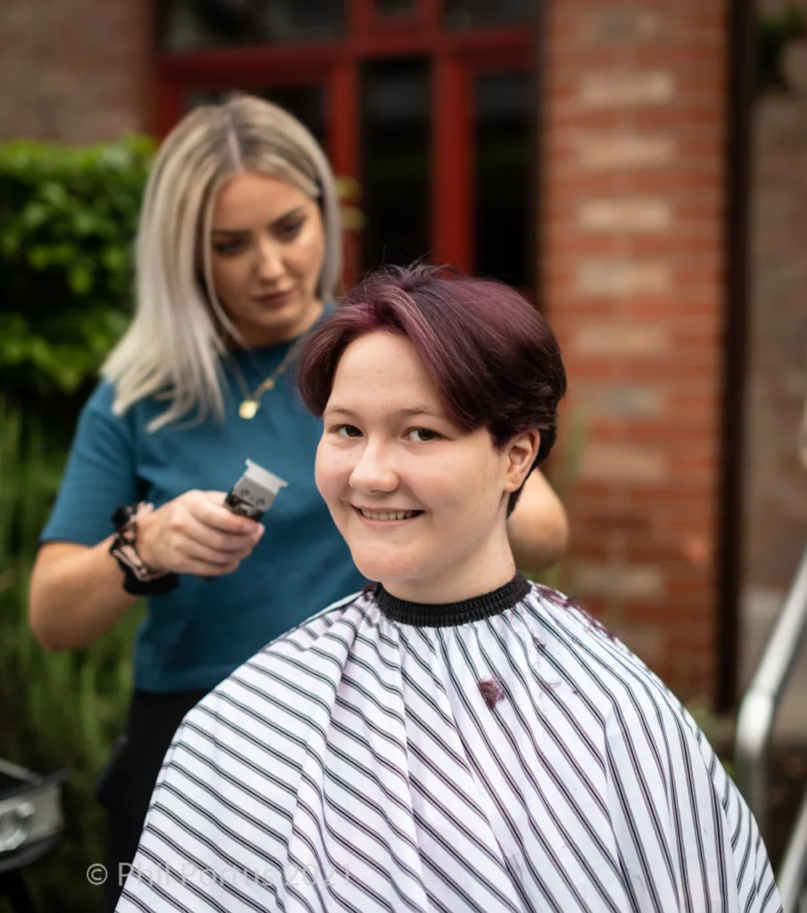 A person with a short, dark purple haircut smiles while a stylist uses clippers nearby.