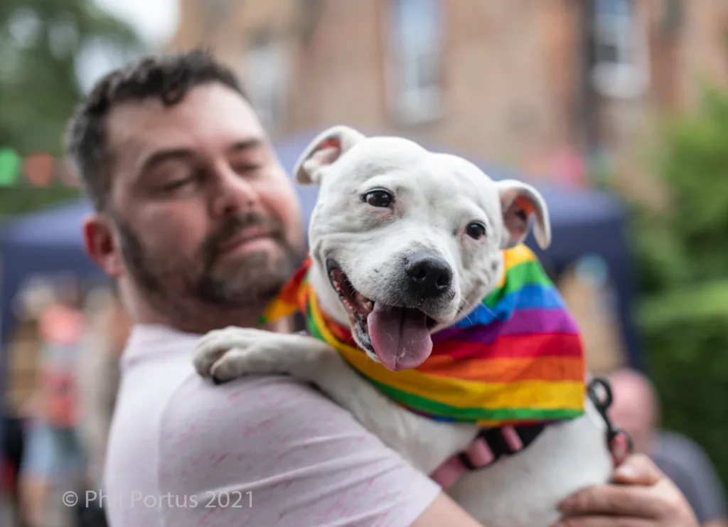 A man holds a white dog wearing a rainbow scarf in his arms.