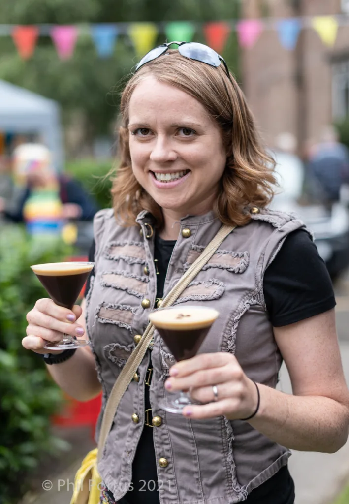 A smiling woman holds two small glasses of a dark liquid while wearing sunglasses and a vest.