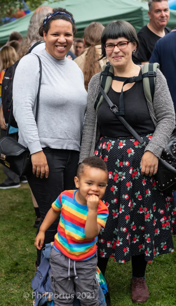Three people stand outdoors on grass; two adults smile with a young child who makes a face.