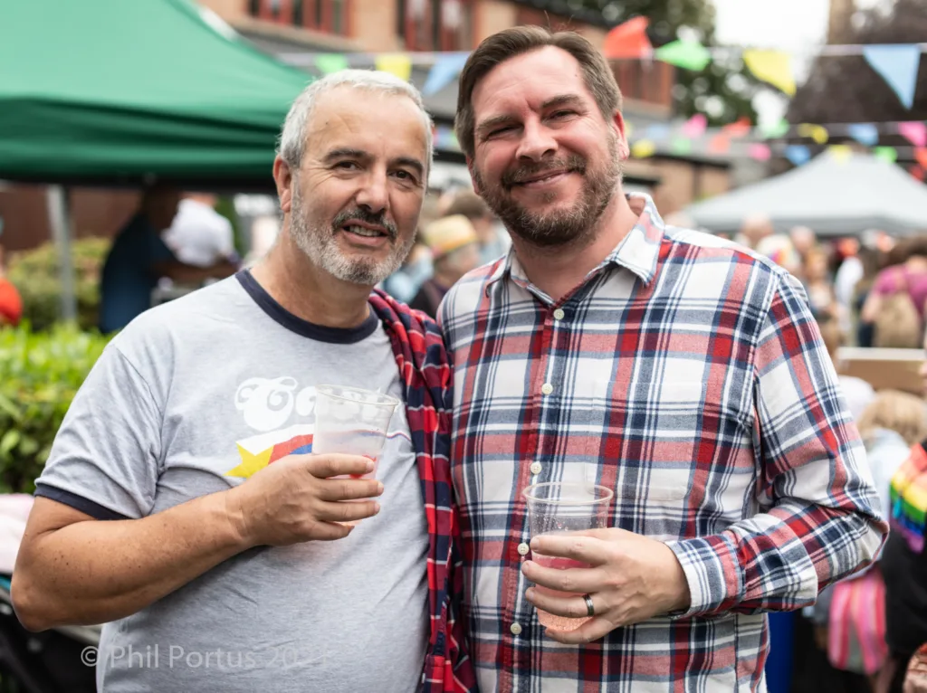 Two men smile while standing close together at an outdoor event, each holding a clear plastic cup.