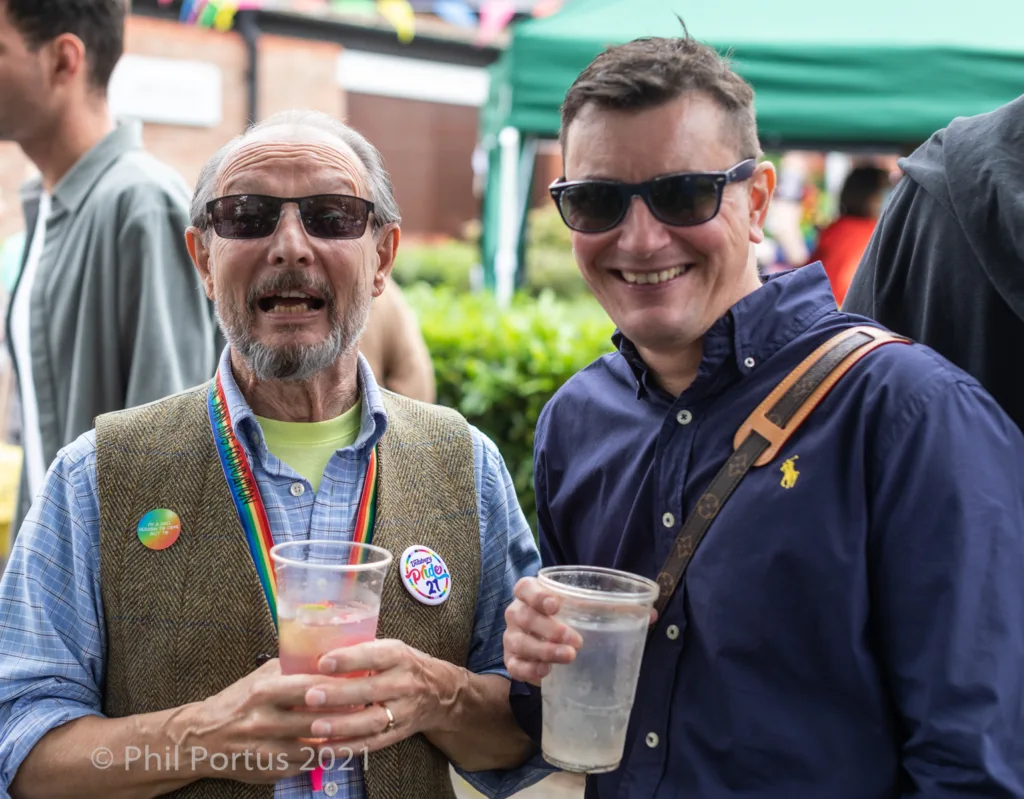Two men wearing sunglasses smile while holding clear plastic cups at an outdoor event.
