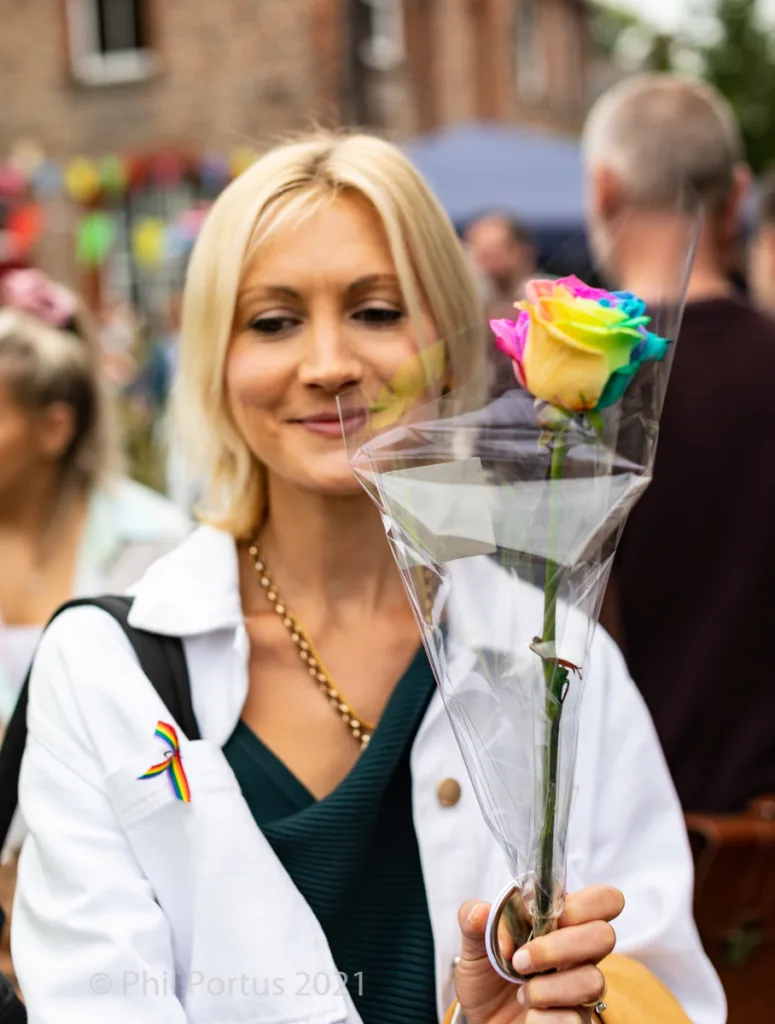 A person with blonde hair holds a rainbow rose in clear plastic wrapping.