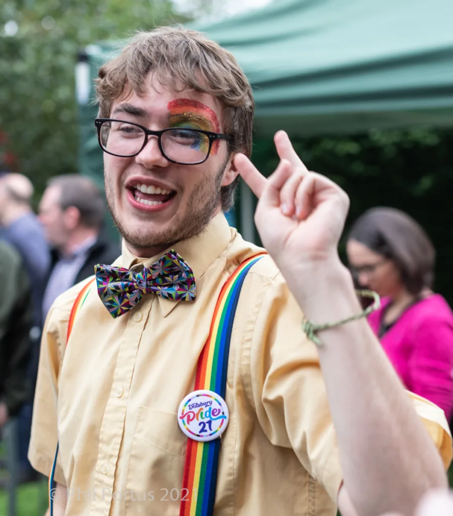 A person with fair skin smiles widely while making a peace sign gesture at an outdoor event.