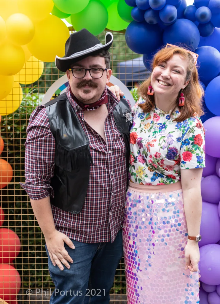 Two people stand close together smiling, wearing costumes amidst colorful balloon decorations.