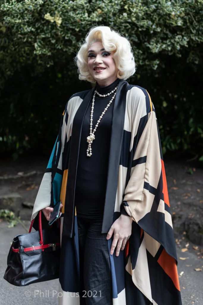 A person with styled blonde hair stands outdoors wearing a black turtleneck and patterned wrap.