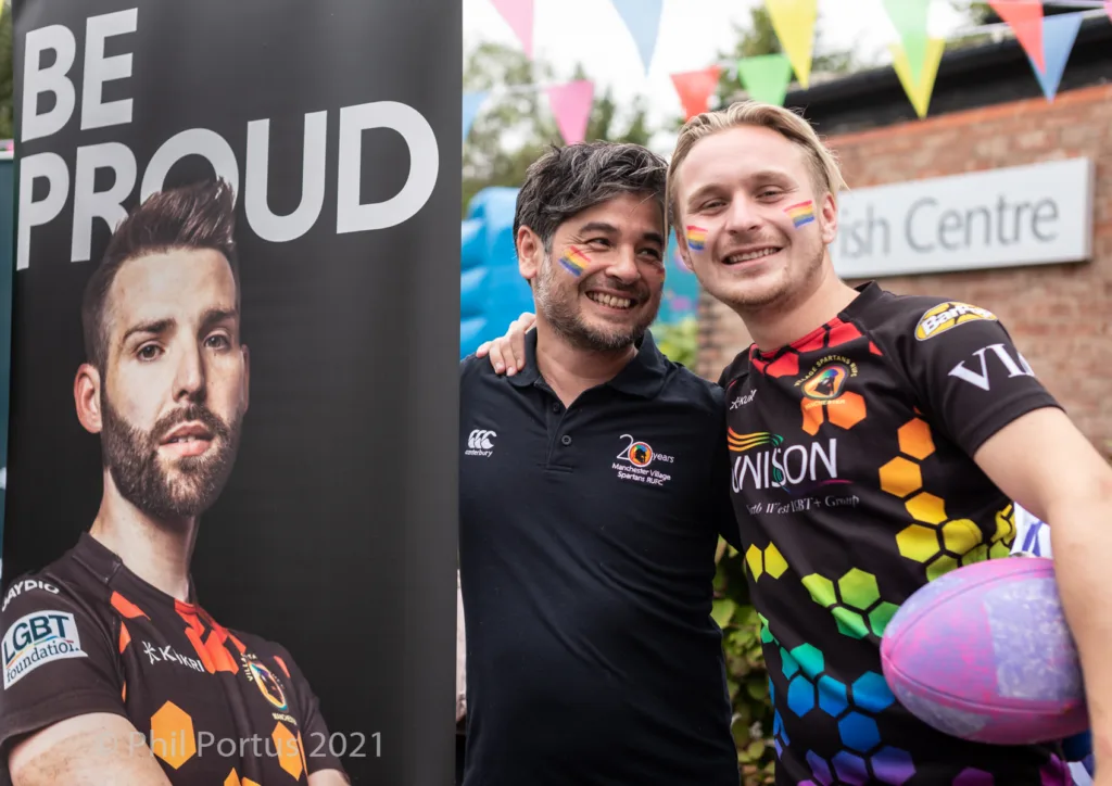 Two men pose with a rugby ball in front of a banner and building decorated with colorful flags.