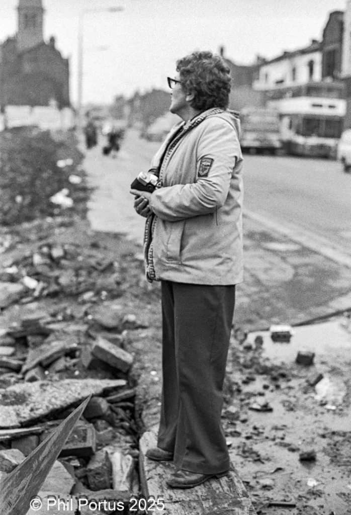 A person with glasses stands near rubble, holding a bottle in their hand.